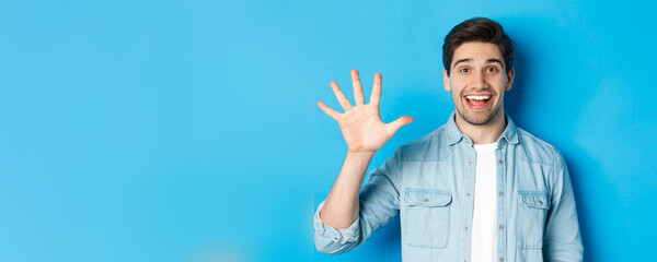 Fototapeta na wymiar Close-up of handsome man smiling, showing fingers number five, standing over blue background