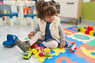 Adorable hispanic toddler playing with car toy sitting on floor at kindergarten