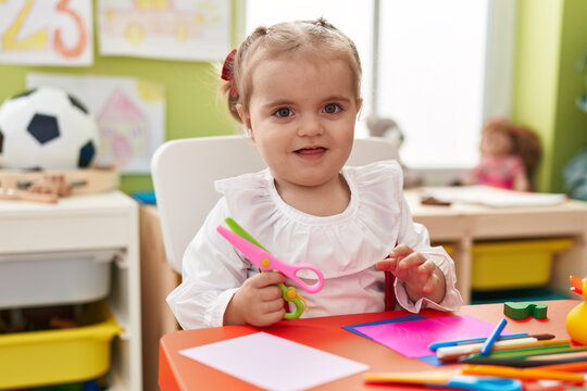 Adorable blonde toddler student sitting on table holding scissors at kindergarten