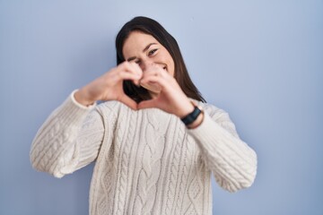 Young brunette woman standing over blue background smiling in love doing heart symbol shape with hands. romantic concept.