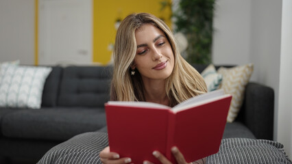 Young blonde woman reading book lying on sofa at home