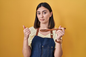 Hispanic girl eating healthy  wooden spoon pointing with hand finger to the side showing advertisement, serious and calm face