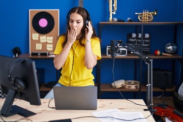 Young hispanic girl listening to music wearing headphones covering mouth with hand, shocked and...