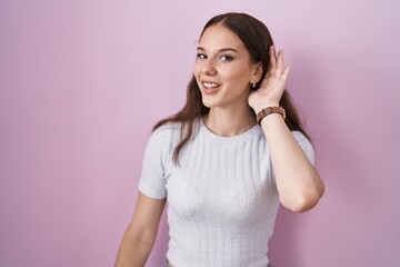 Young hispanic girl standing over pink background smiling with hand over ear listening an hearing to rumor or gossip. deafness concept.