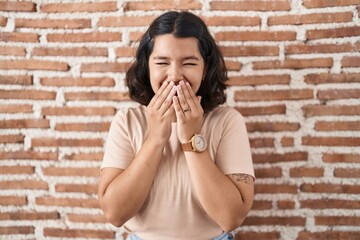 Young hispanic woman standing over bricks wall laughing and embarrassed giggle covering mouth with...