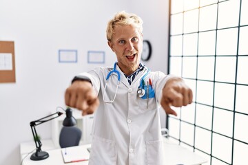 Young blond man wearing doctor uniform and stethoscope at clinic pointing to you and the camera with fingers, smiling positive and cheerful