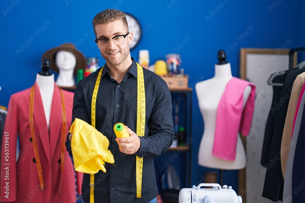 Wall mural Young caucasian man tailor smiling confident holding cloth and thread at clothing factory