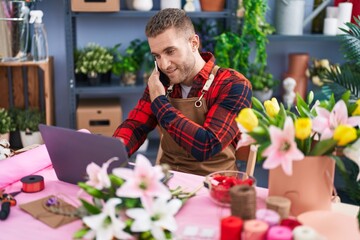 Young caucasian man florist talking on smartphone using laptop at flower shop