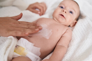 Adorable caucasian baby lying on bed applying cream on body skin at bedroom