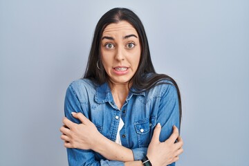 Hispanic woman standing over blue background shaking and freezing for winter cold with sad and...