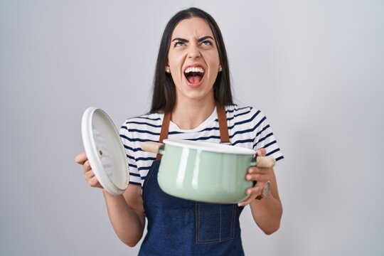 Young brunette woman wearing apron holding cooking pot angry and mad screaming frustrated and furious, shouting with anger looking up.