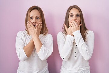 Middle age mother and young daughter standing over pink background shocked covering mouth with hands for mistake. secret concept.
