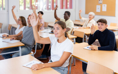 Young positive woman student sitting at desk and raising her hand to answer teacher's question in classroom with group of people.