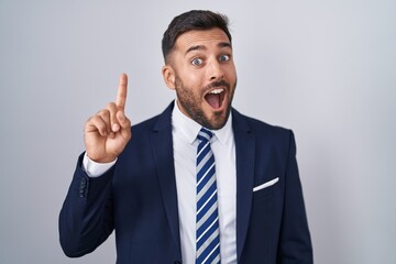 Handsome hispanic man wearing suit and tie pointing finger up with successful idea. exited and happy. number one.