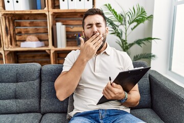 Handsome hispanic man holding clipboard working at psychology clinic bored yawning tired covering...