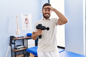 Naklejka na ściany i meble Handsome hispanic man holding therapy massage gun at physiotherapy center stressed and frustrated with hand on head, surprised and angry face