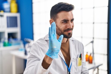 Young hispanic man wearing scientist uniform saying hello with hand at laboratory