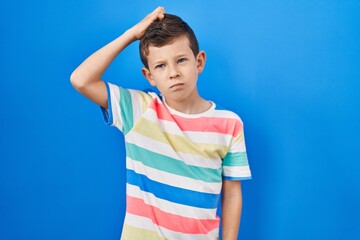 Young caucasian kid standing over blue background confuse and wondering about question. uncertain with doubt, thinking with hand on head. pensive concept.