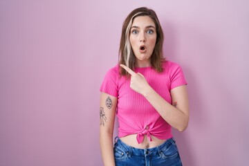 Blonde caucasian woman standing over pink background surprised pointing with finger to the side, open mouth amazed expression.