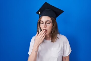 Blonde caucasian woman wearing graduation cap bored yawning tired covering mouth with hand. restless and sleepiness.
