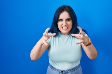 Fototapeta premium Young modern girl with blue hair standing over blue background shouting frustrated with rage, hands trying to strangle, yelling mad