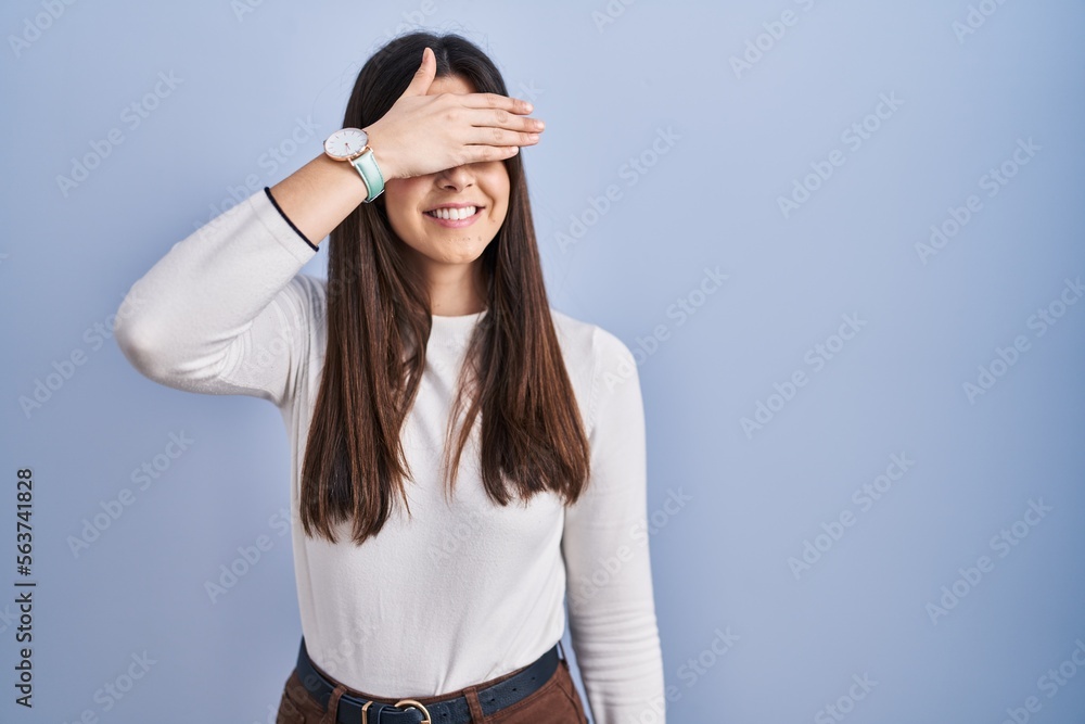 Poster Young brunette woman standing over blue background smiling and laughing with hand on face covering eyes for surprise. blind concept.