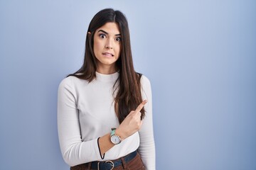 Young brunette woman standing over blue background pointing aside worried and nervous with...