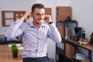 Young hispanic man at the office smiling pulling ears with fingers, funny gesture. audition problem