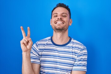 Young hispanic man standing over blue background smiling with happy face winking at the camera doing victory sign with fingers. number two.