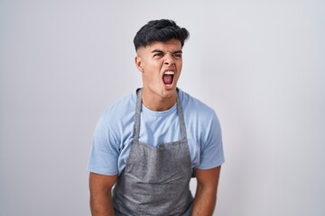 Fototapeta premium Hispanic young man wearing apron over white background angry and mad screaming frustrated and furious, shouting with anger. rage and aggressive concept.