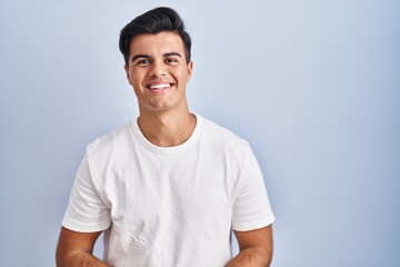 Hispanic man standing over blue background with hands together and crossed fingers smiling relaxed and cheerful. success and optimistic
