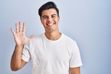 Hispanic man standing over blue background showing and pointing up with fingers number five while smiling confident and happy.