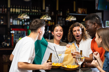 Group of excited young adults of different nationalities, sports fans celebrating victory of favorite team with beer in bar, waving national flag of Ireland