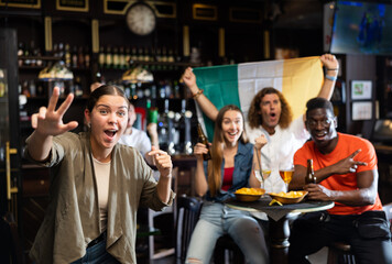 Excited young woman, Ireland football team fan, gesturing with hands, spending time in bar with friends. People with state flag in pub.