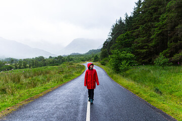 Portrait of a woman wearing a red raincoat while walking alone on a road that crosses a valley surrounded by mountains and fir trees during a foggy day in the highlands, Scotland