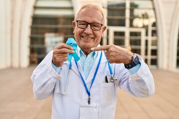 Senior doctor with grey hair holding blue ribbon smiling happy pointing with hand and finger