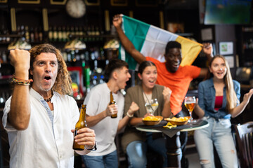 Happy adult football fan drinking beer and watching game on TV in sports bar proud of favorite team victory on background of group of cheerful people with national flag of Ireland