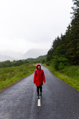 Portrait of a woman wearing a red raincoat while walking alone on a road that crosses a valley surrounded by mountains and fir trees during a foggy day in the highlands, Scotland