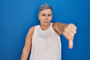 Fototapeta premium Middle age caucasian woman standing over blue background looking unhappy and angry showing rejection and negative with thumbs down gesture. bad expression.