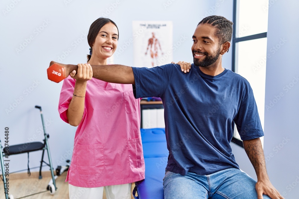 Sticker Man and woman wearing physiotherapist uniform having rehab session stretching arm holding dumbbell at physiotherpy clinic