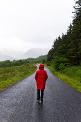 Portrait of a person wearing a red raincoat while walking alone on a road that crosses a valley surrounded by mountains and fir trees during a foggy day in the highlands, Scotland