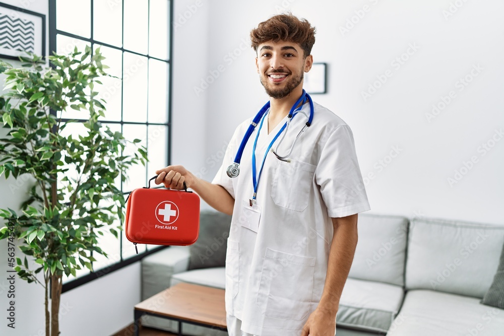 Sticker young arab man wearing doctor uniform holding first aid box at clinic