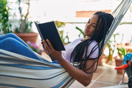 African american woman reading book lying on hammock at home terrace
