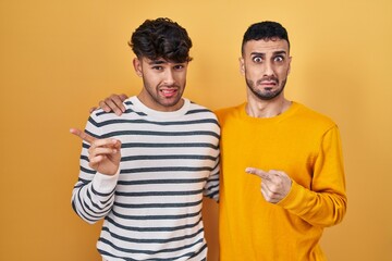 Young hispanic gay couple standing over yellow background pointing aside worried and nervous with forefinger, concerned and surprised expression
