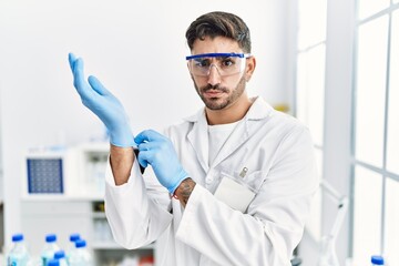 Young hispanic man working at scientist laboratory putting gloves on skeptic and nervous, frowning...