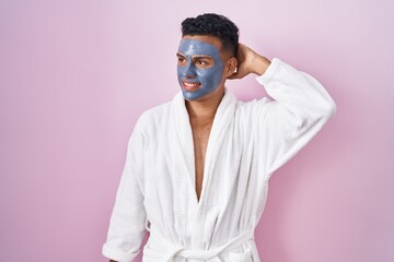 Young hispanic man wearing beauty face mask and bath robe smiling confident touching hair with hand up gesture, posing attractive and fashionable