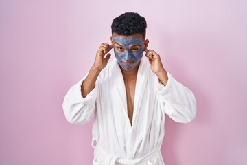Young hispanic man wearing beauty face mask and bath robe covering ears with fingers with annoyed expression for the noise of loud music. deaf concept.