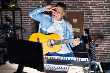 Hispanic young man playing classic guitar at music studio doing peace symbol with fingers over face, smiling cheerful showing victory
