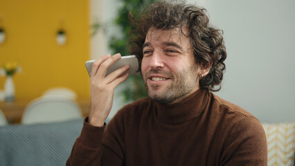 Young hispanic man listening voice message by smartphone sitting on sofa at home