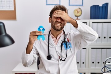 Young hispanic doctor man holding blue ribbon stressed and frustrated with hand on head, surprised and angry face